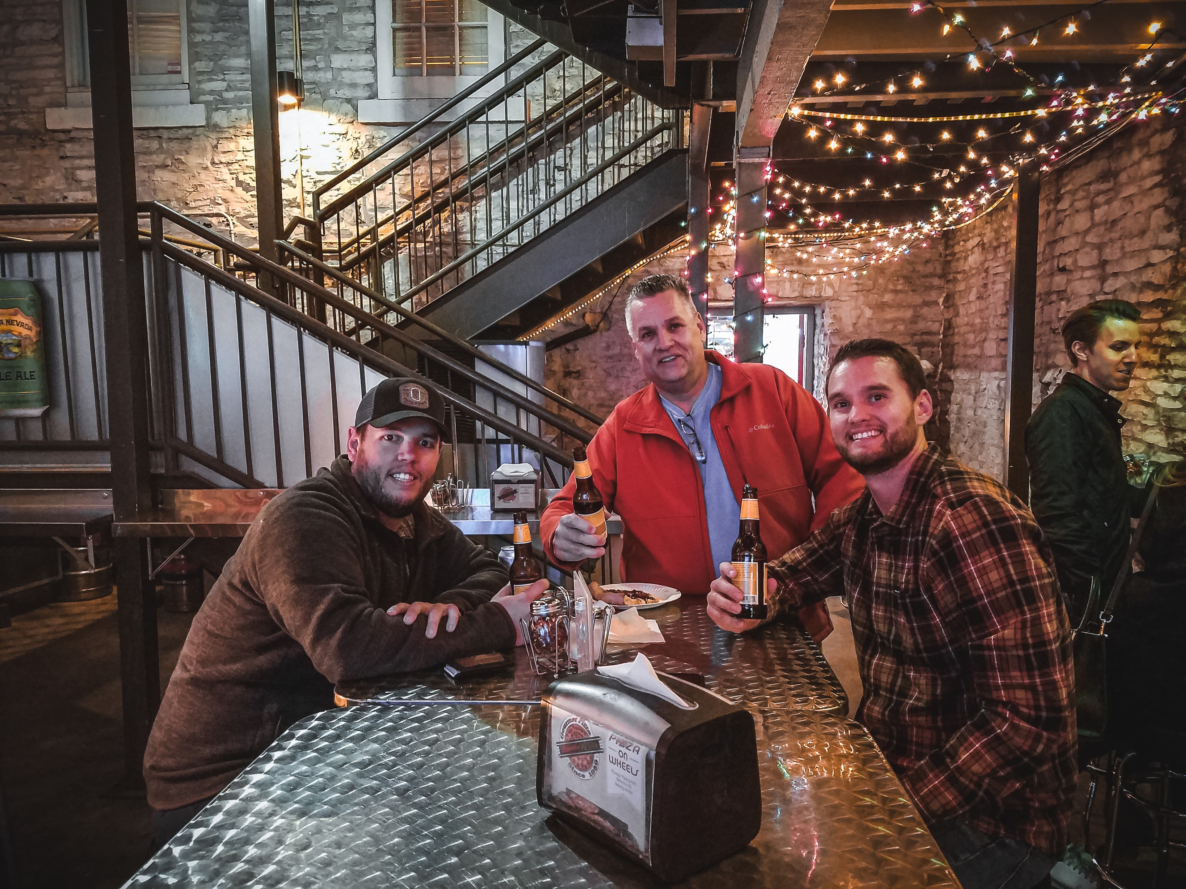 Three men holding beer bottles smile at a metal table under festive string lights.