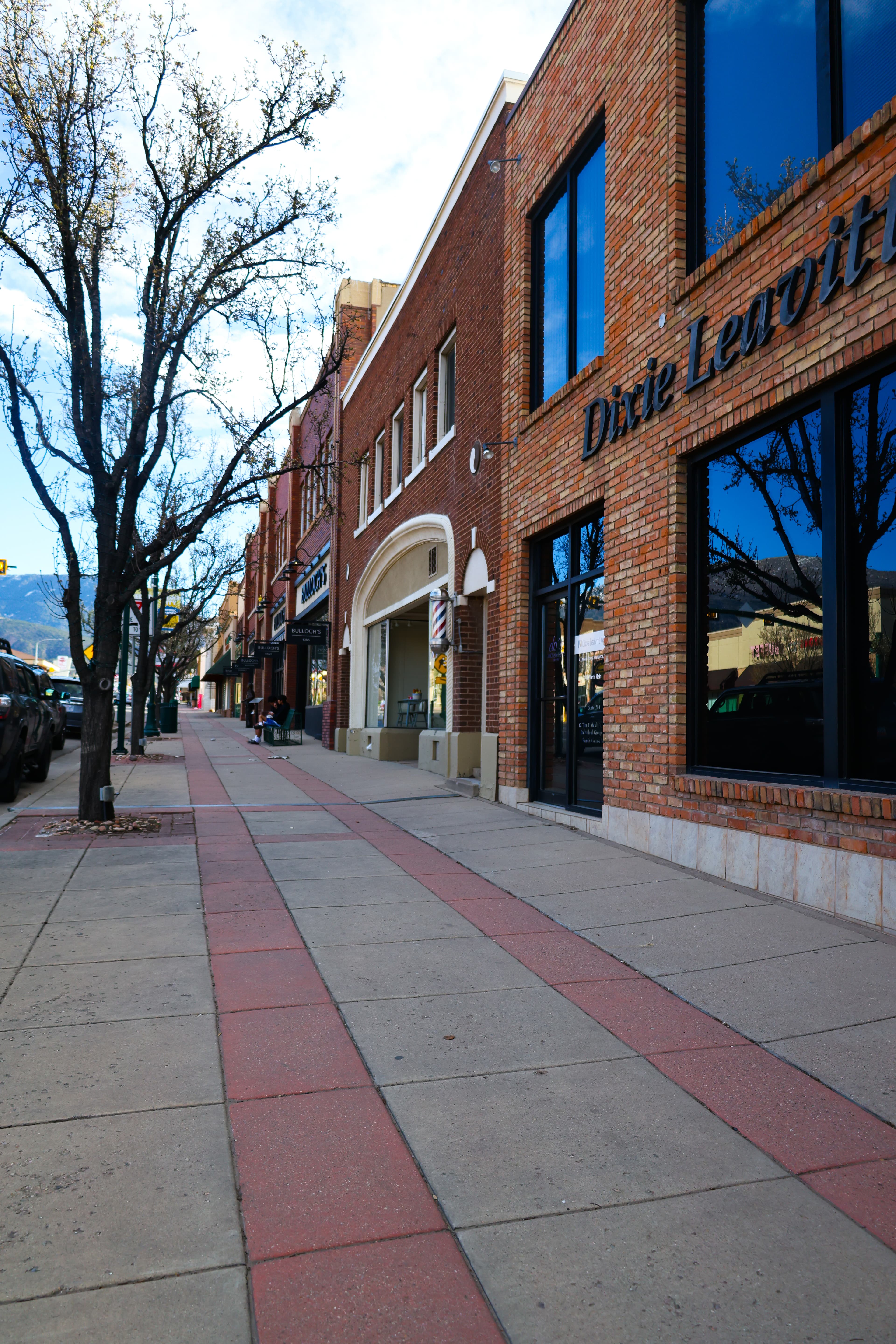 Wide sidewalk with red stripes borders brick storefronts and bare trees in a downtown area.