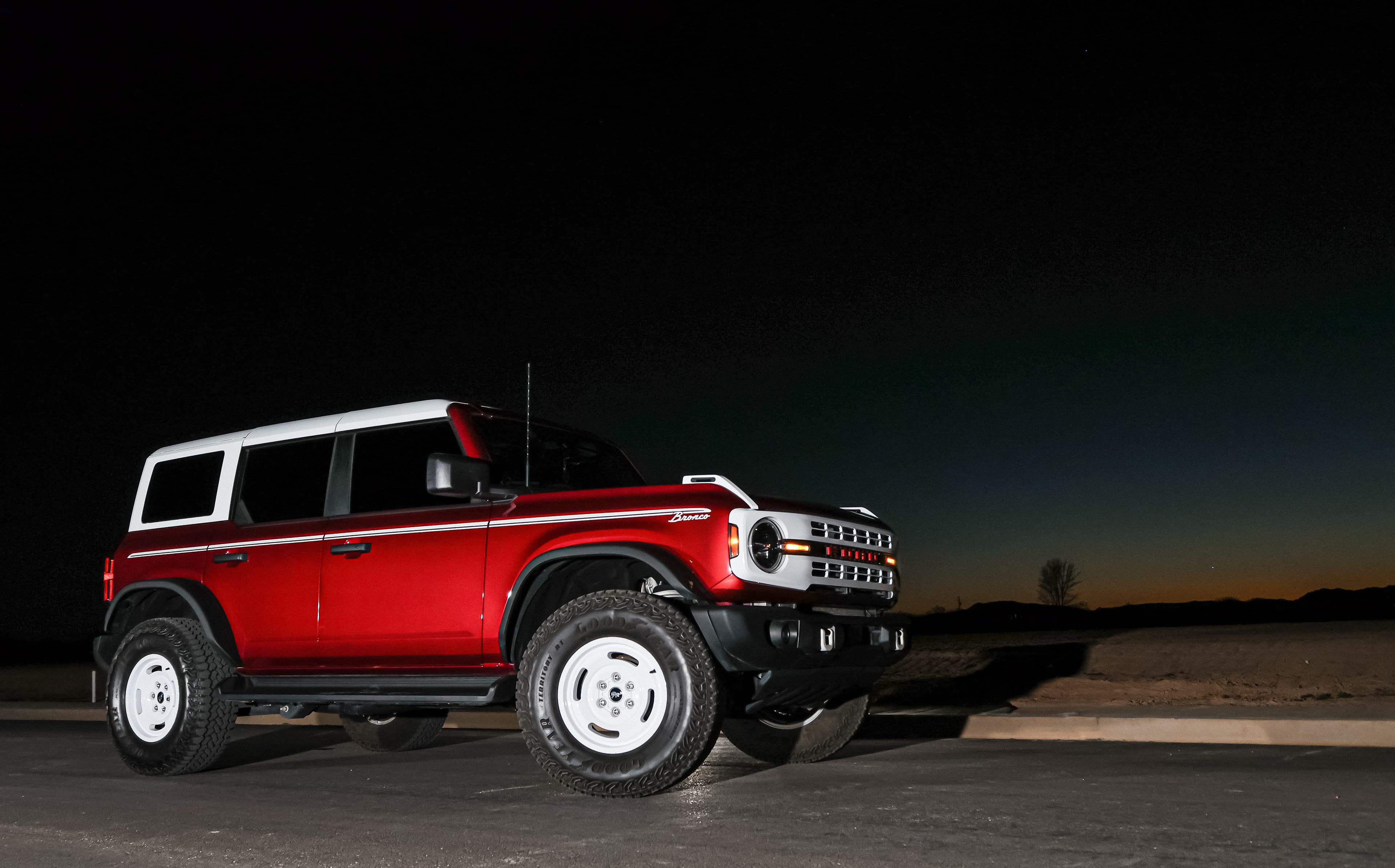 Red Ford Bronco with white roof and wheels parked on a road at night.