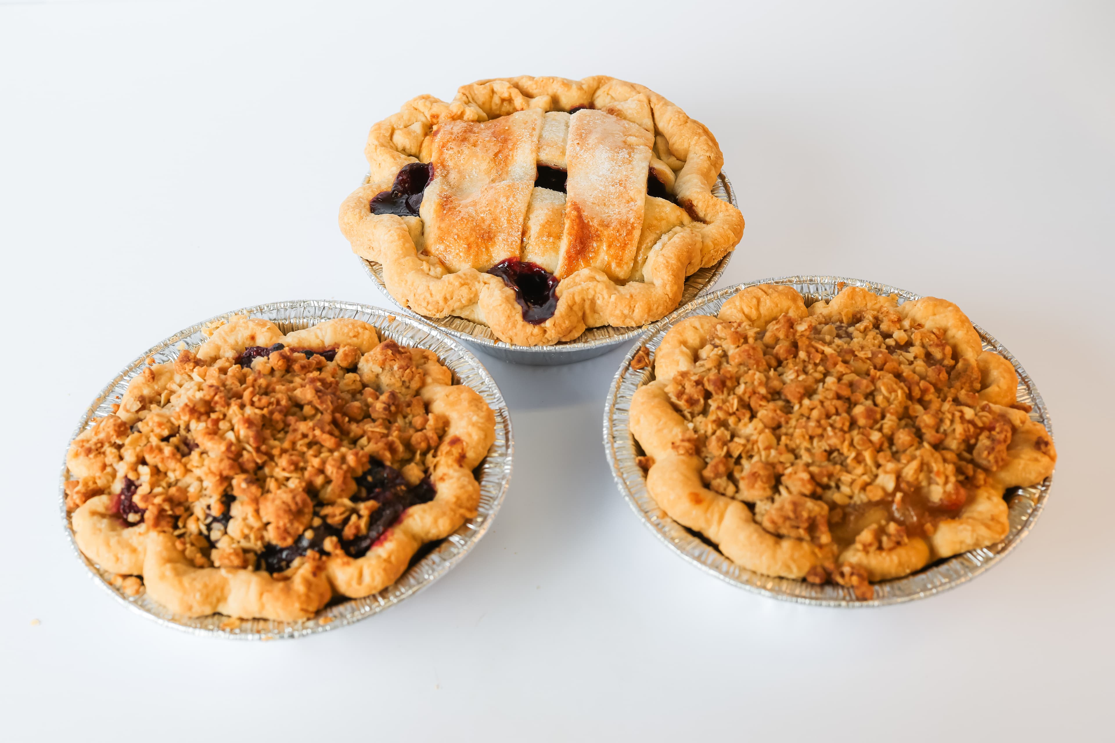 Three small fruit pies in foil tins: one lattice-topped and two with golden crumble toppings.