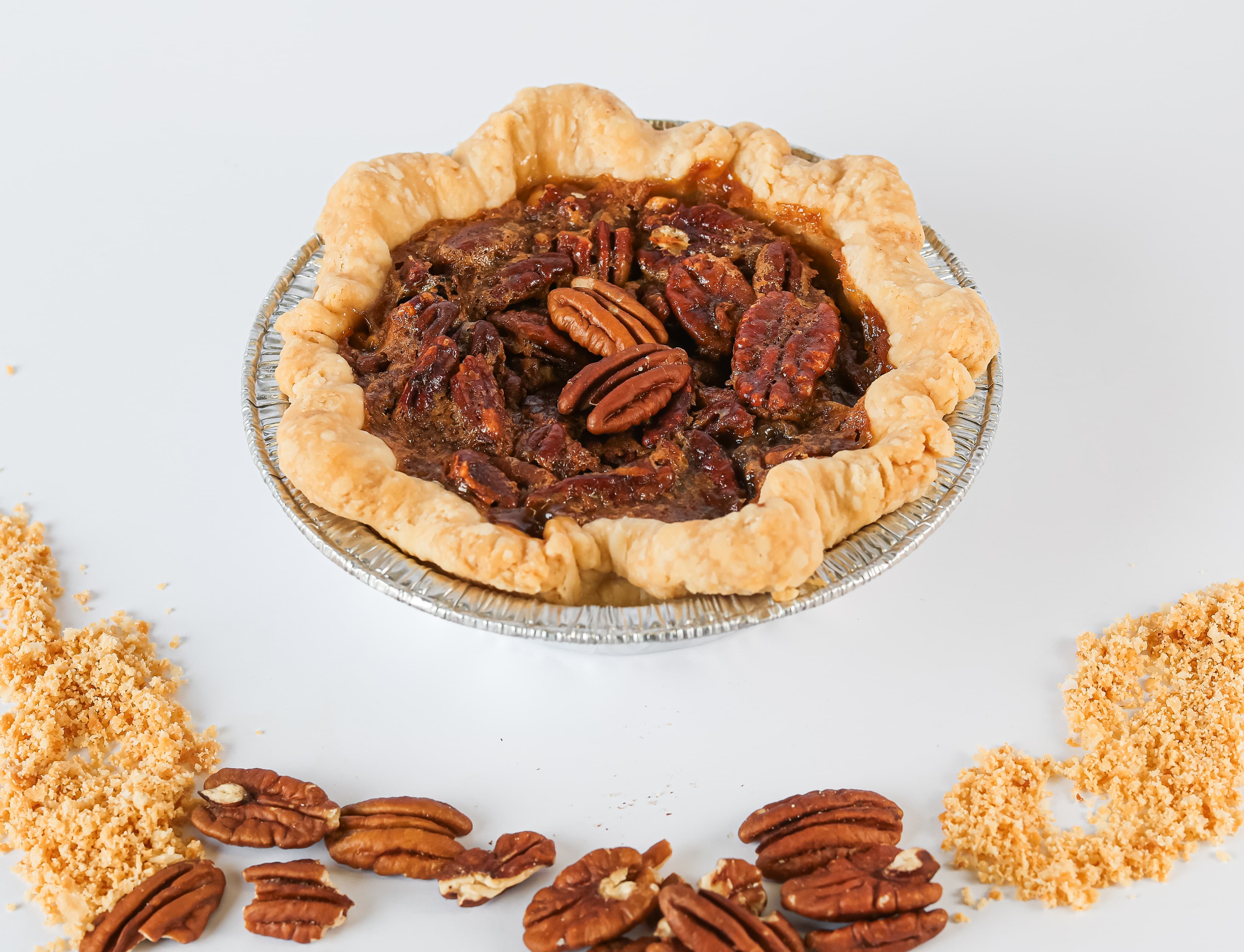 Mini pecan pie with crimped crust and whole pecans, surrounded by scattered nuts and crumbs.