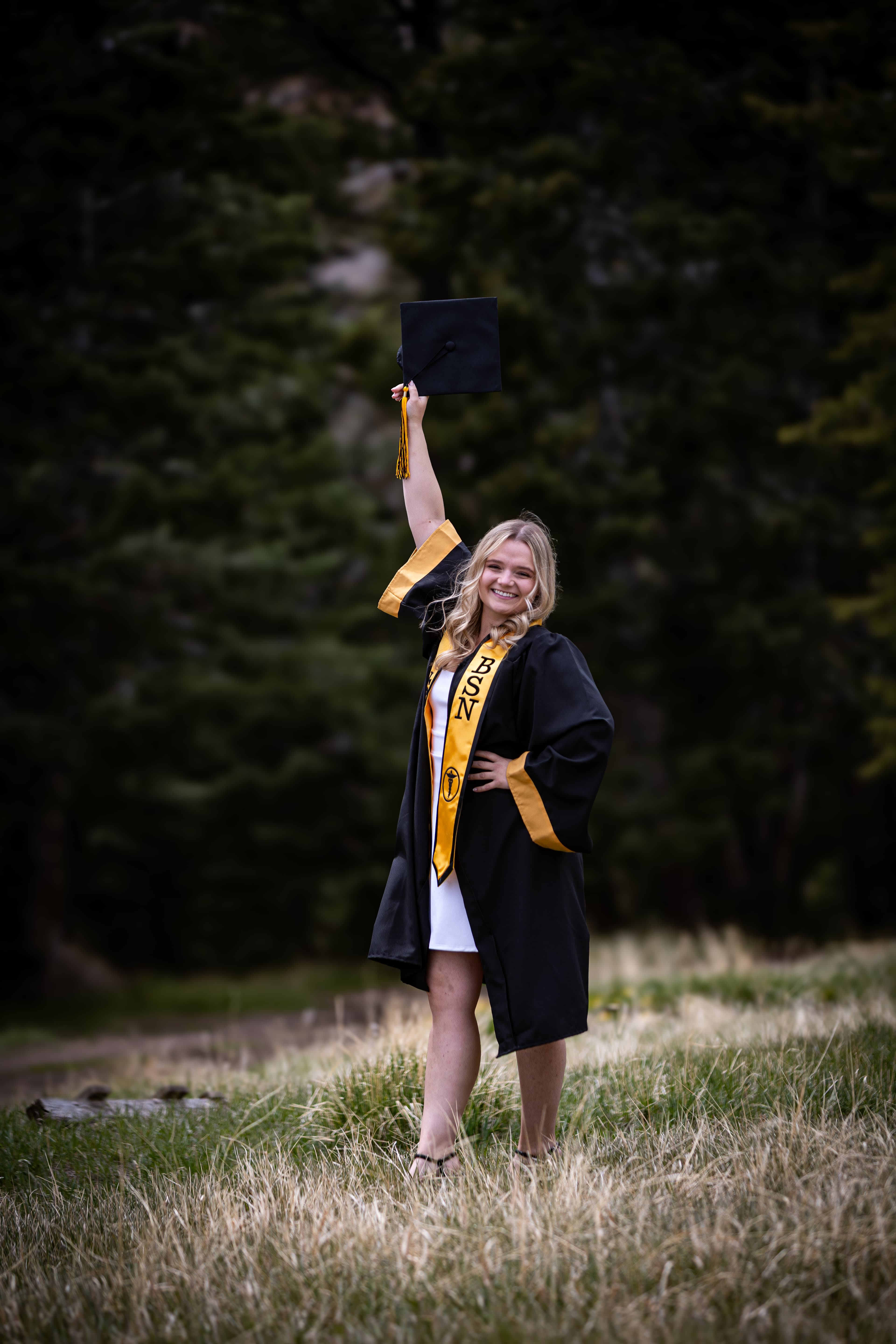 Smiling graduate in a BSN stole holds her cap high in a grassy field.
