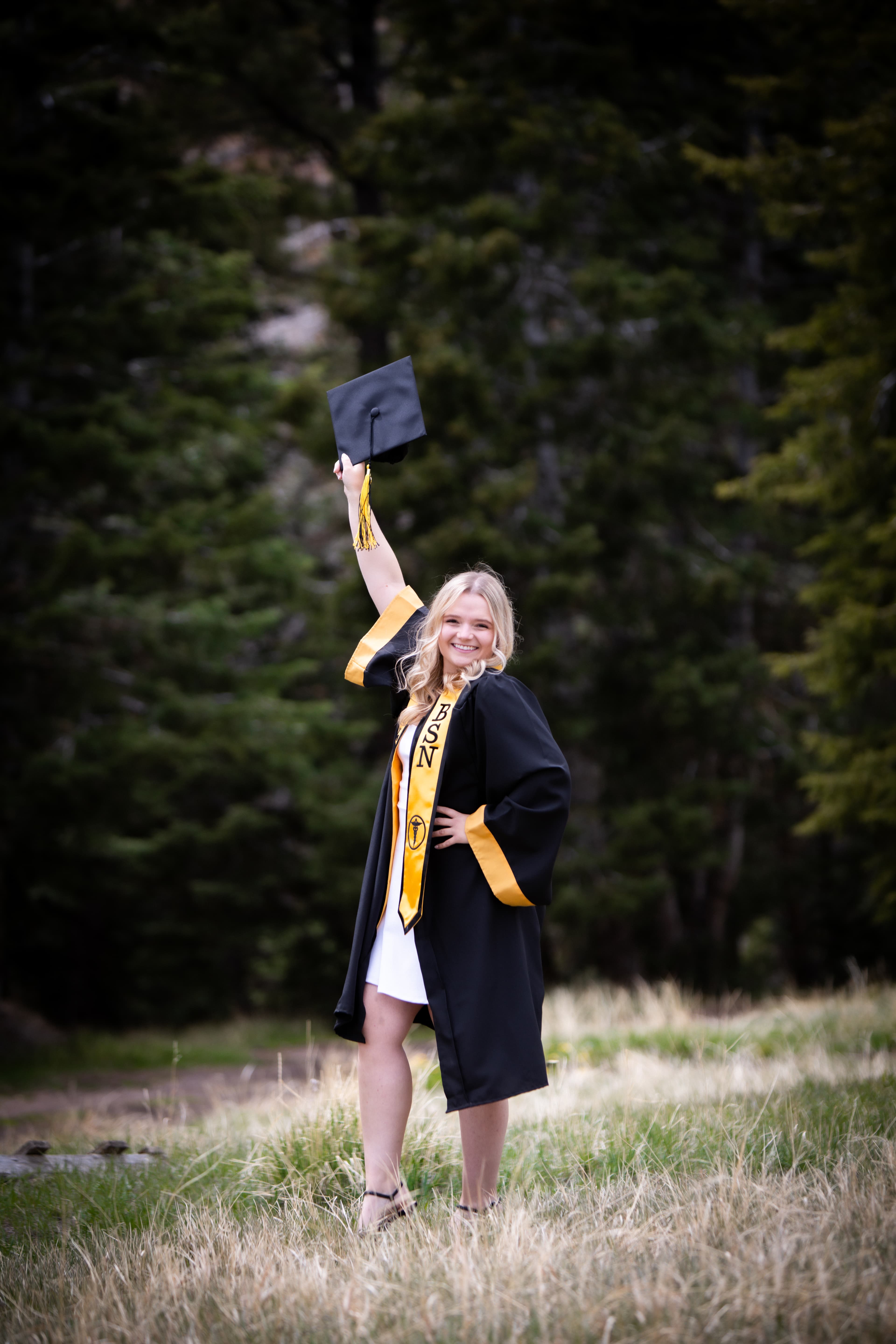 Smiling woman in BSN graduation gown holds her cap high in a grassy field.