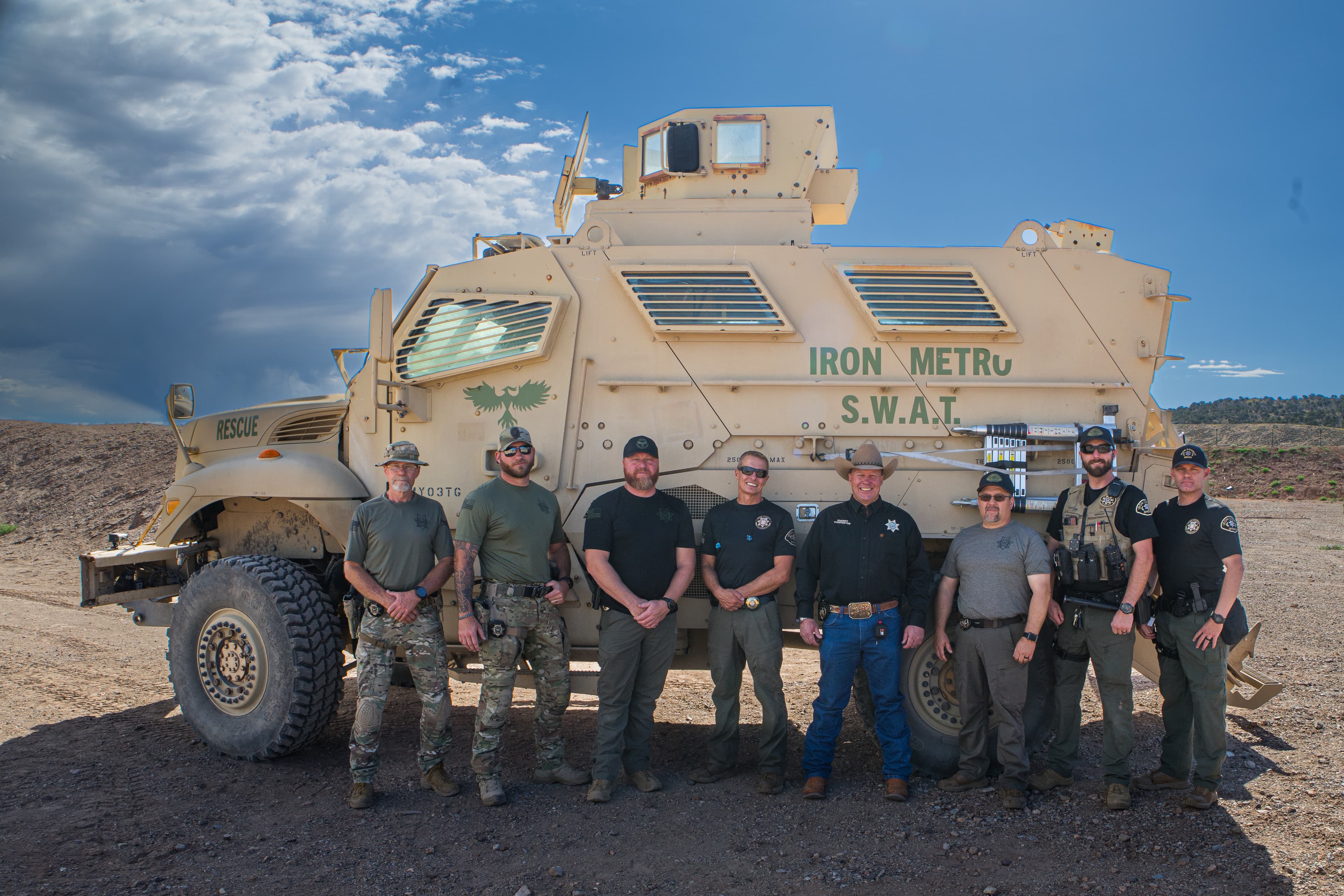 Eight law enforcement officers pose before a tan Iron Metro SWAT armored rescue vehicle.