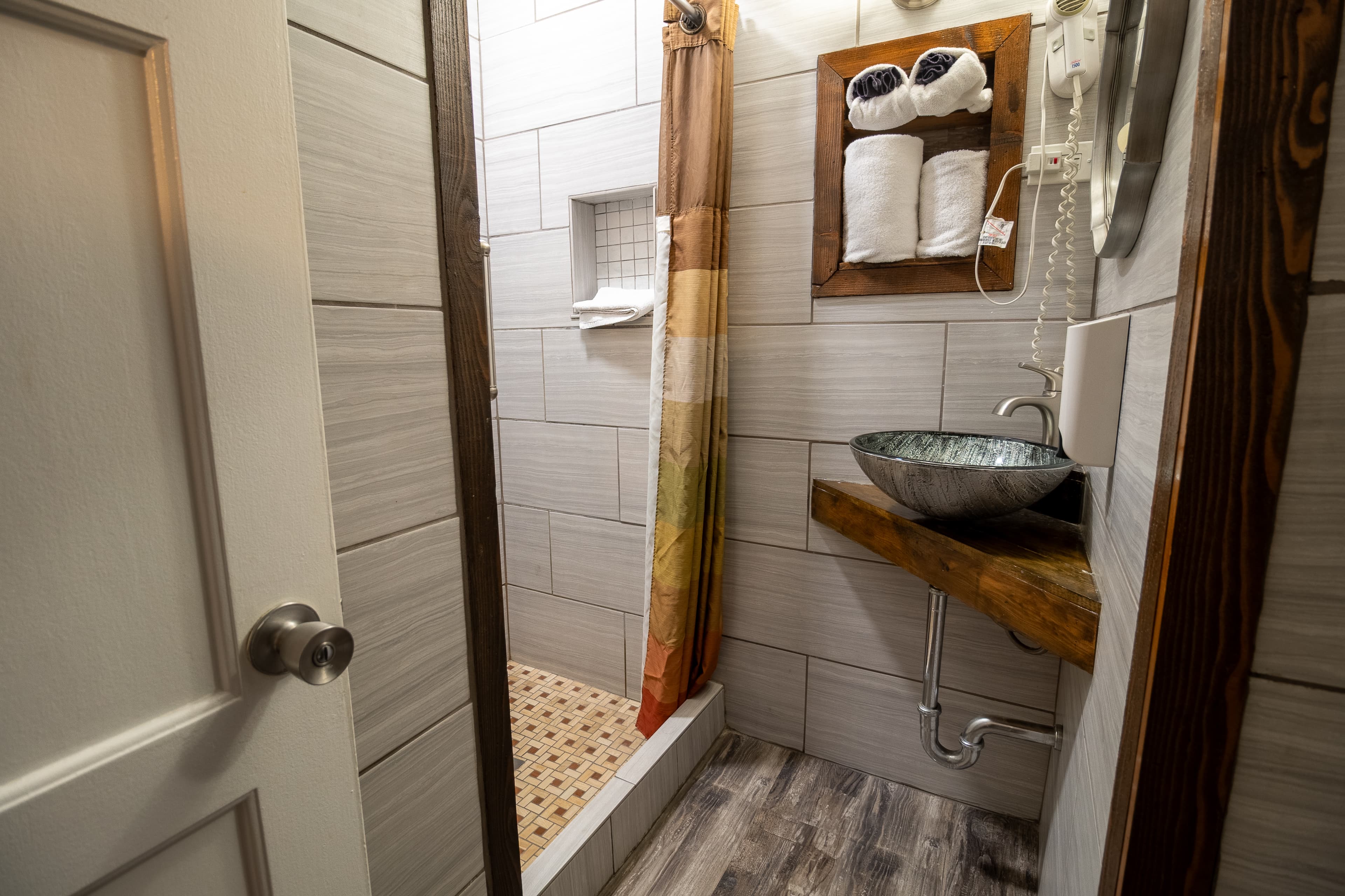 Small bathroom with gray tiles, walk-in shower, and vessel sink on a rustic wooden shelf.