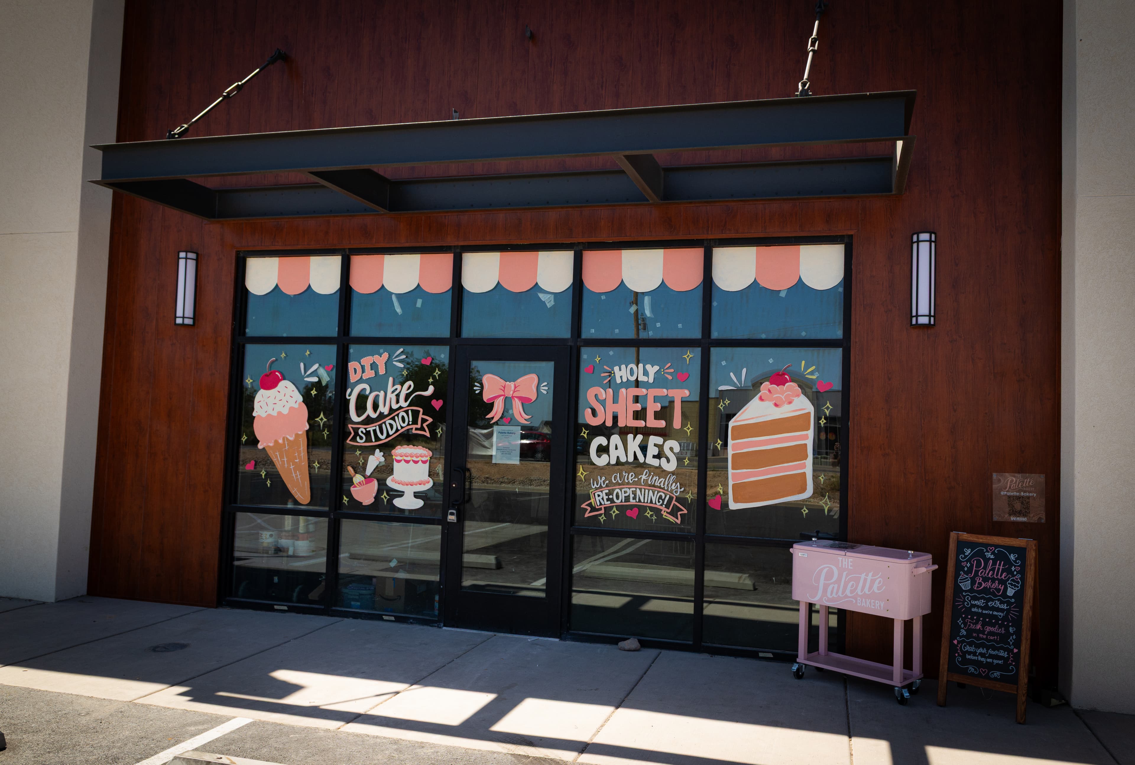 Wood-paneled bakery storefront with colorful window art, a pink cooler, and a chalkboard sign.