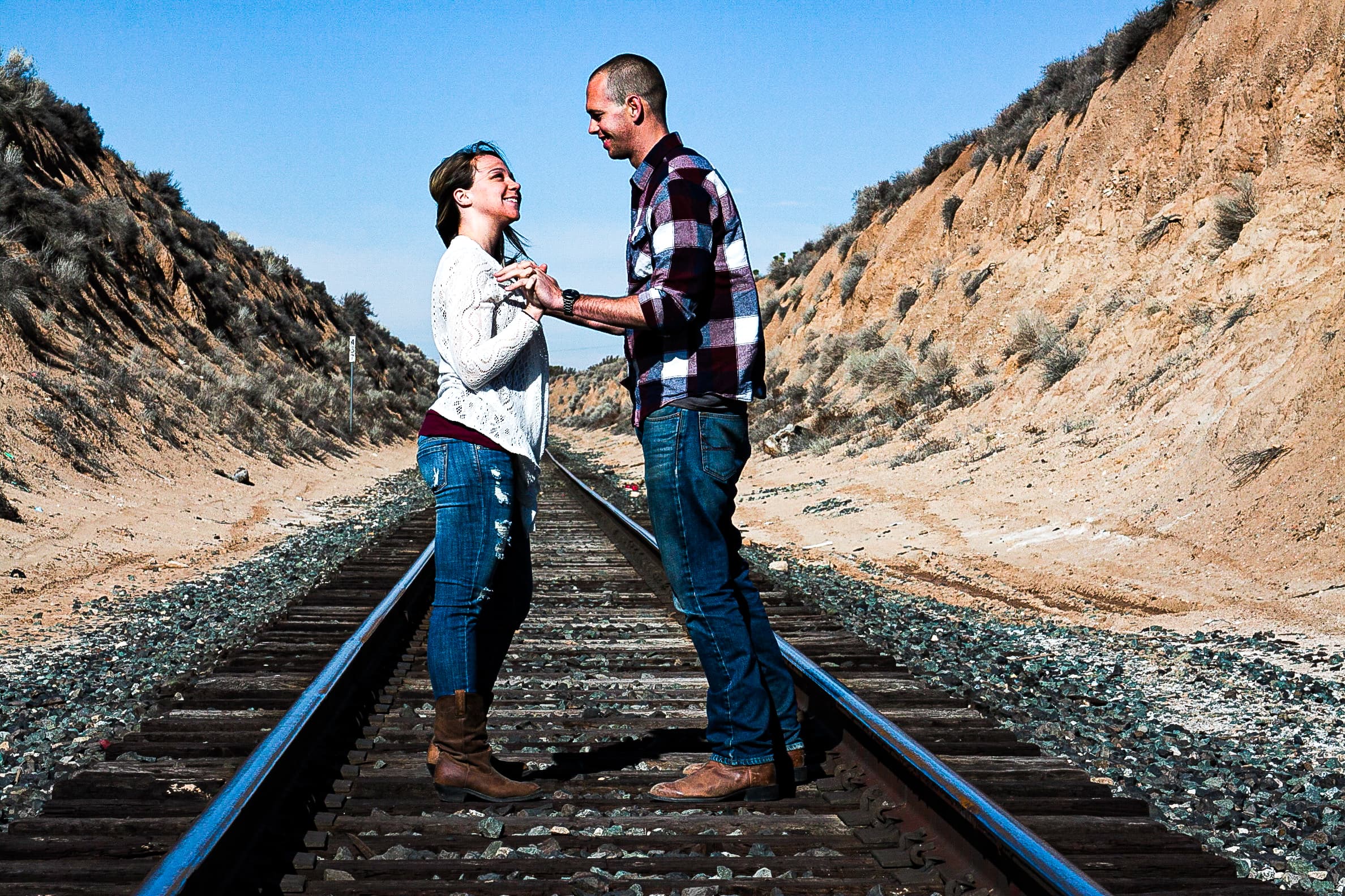 Smiling couple holding hands on a railroad track in a desert canyon under blue skies.
