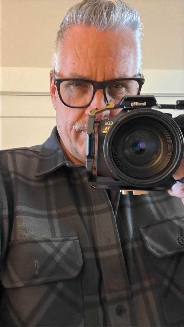 Grey-haired man in a plaid shirt holds a professional camera with a large lens.