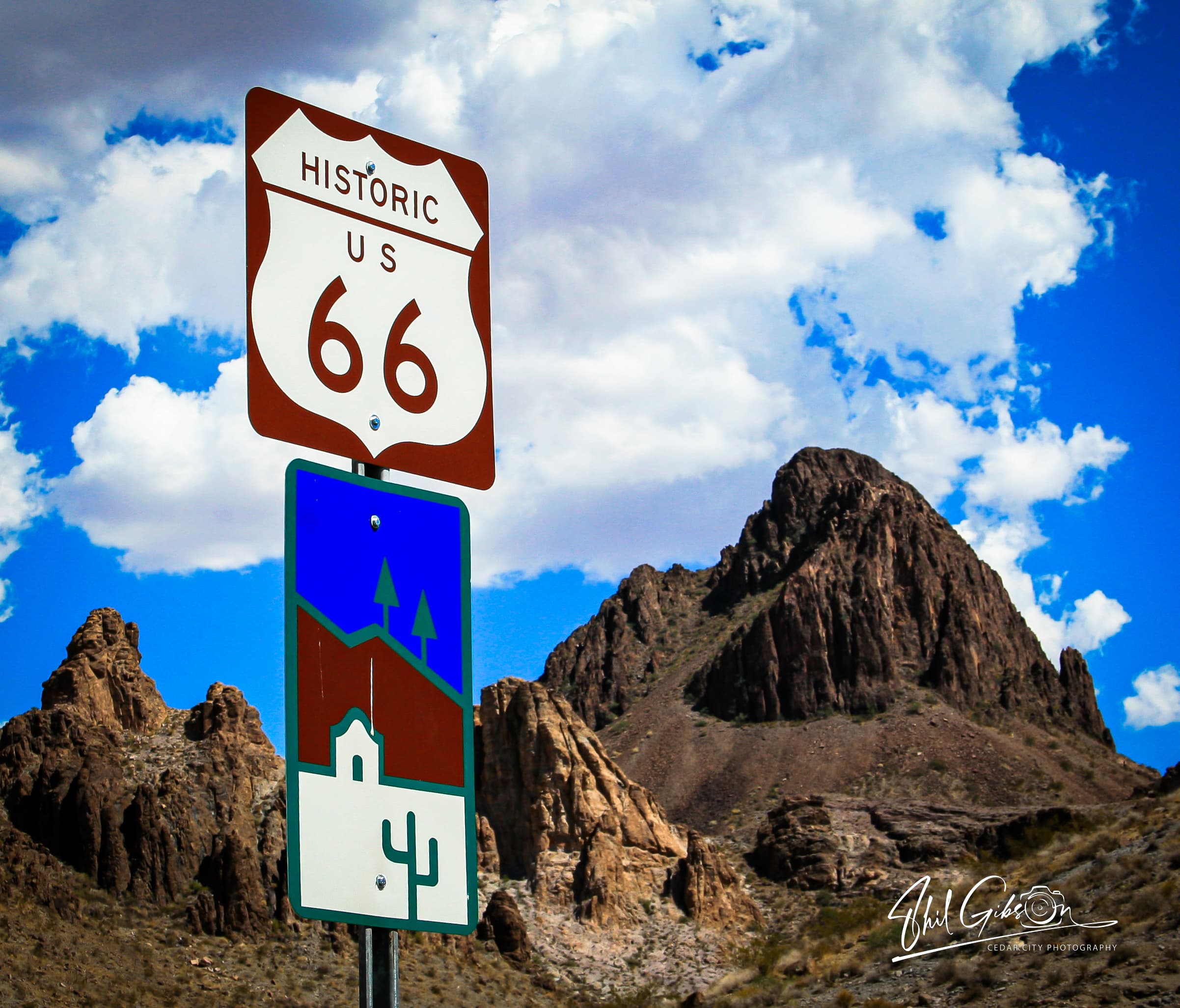 Historic Route 66 sign and colorful desert graphic sign before rugged mountains and blue sky.