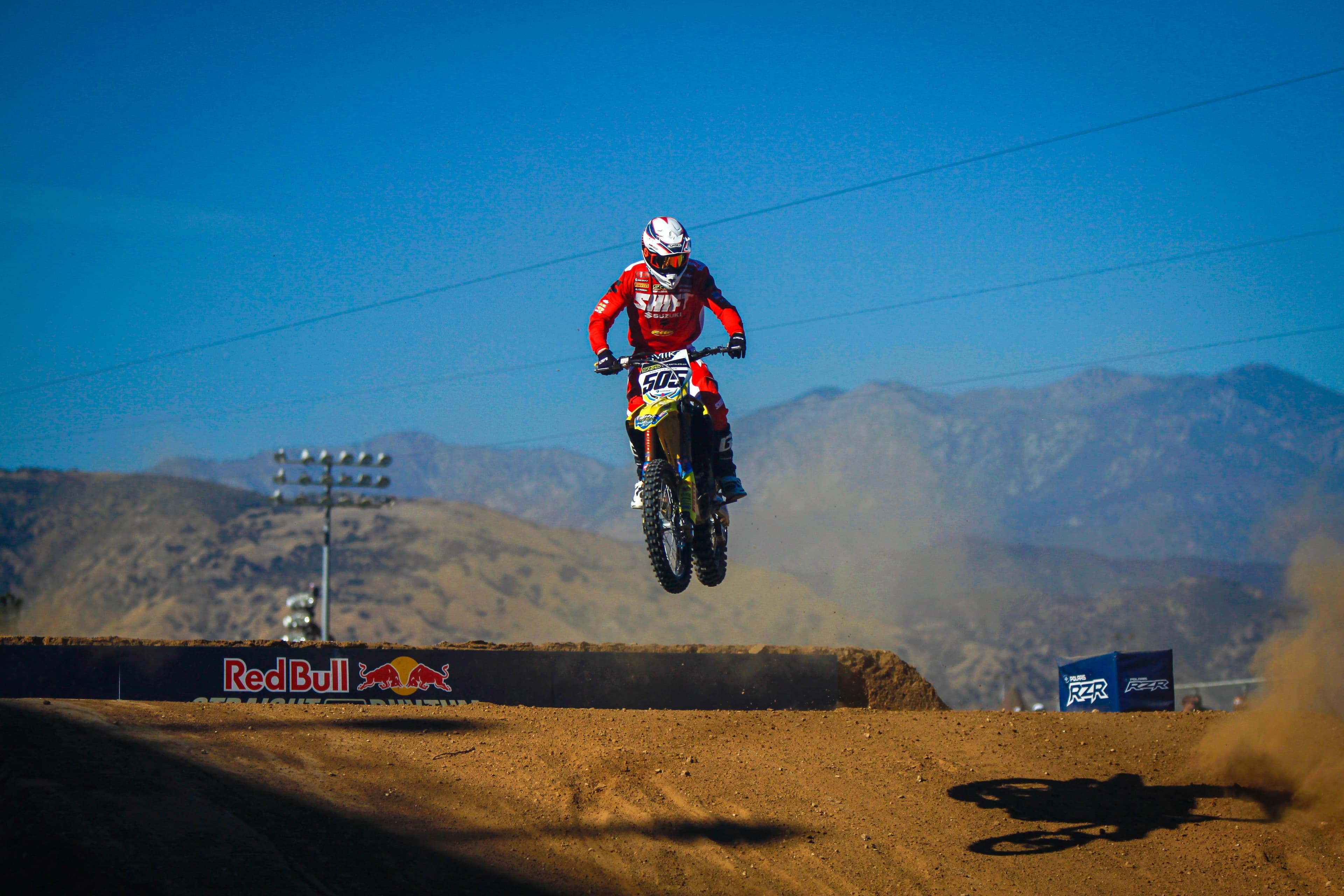Motocross rider in red gear jumps a Suzuki bike over a dirt track against mountains.