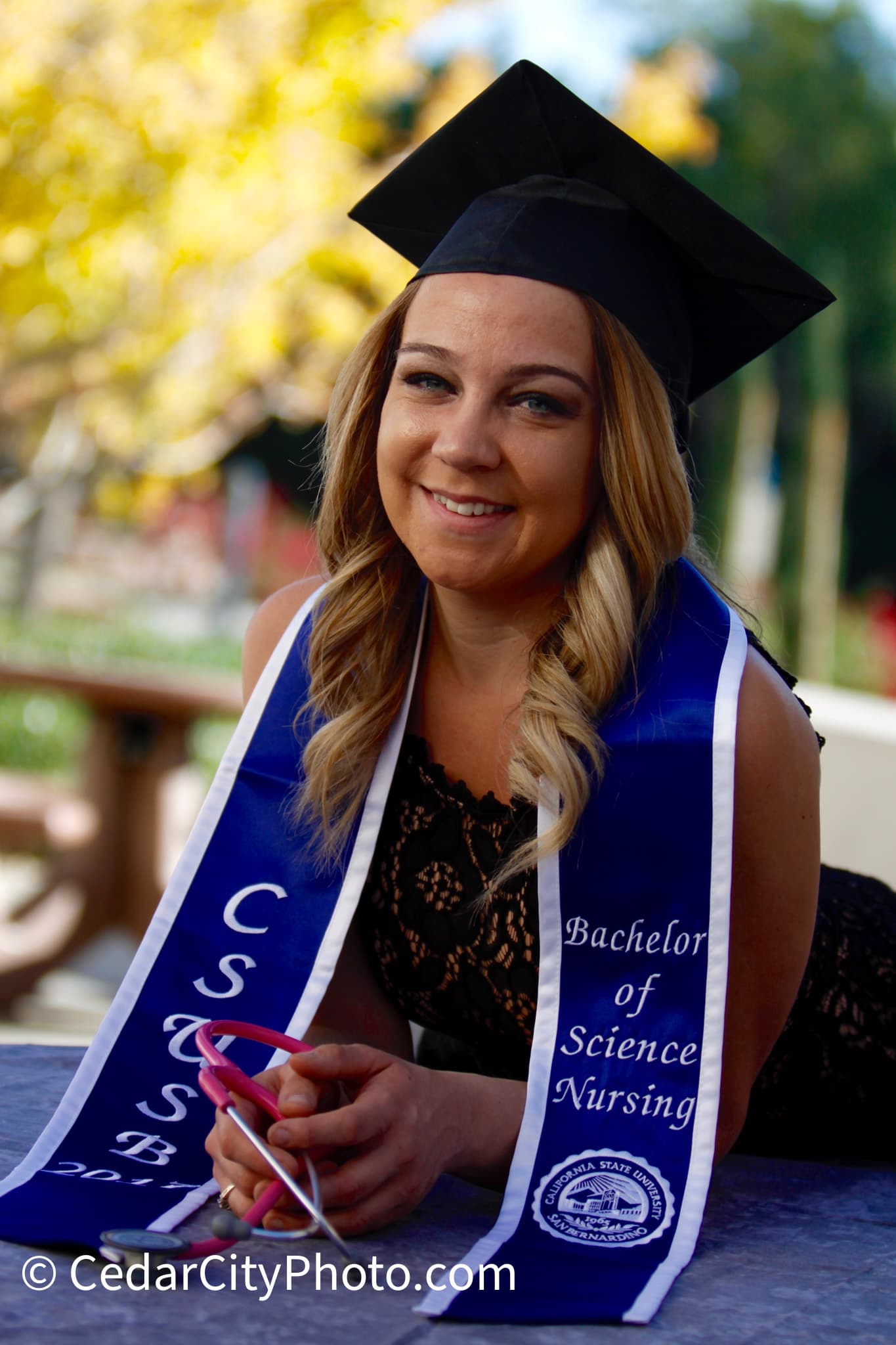 Smiling nursing graduate in cap and CSUSB stole, holding a pink stethoscope outdoors.