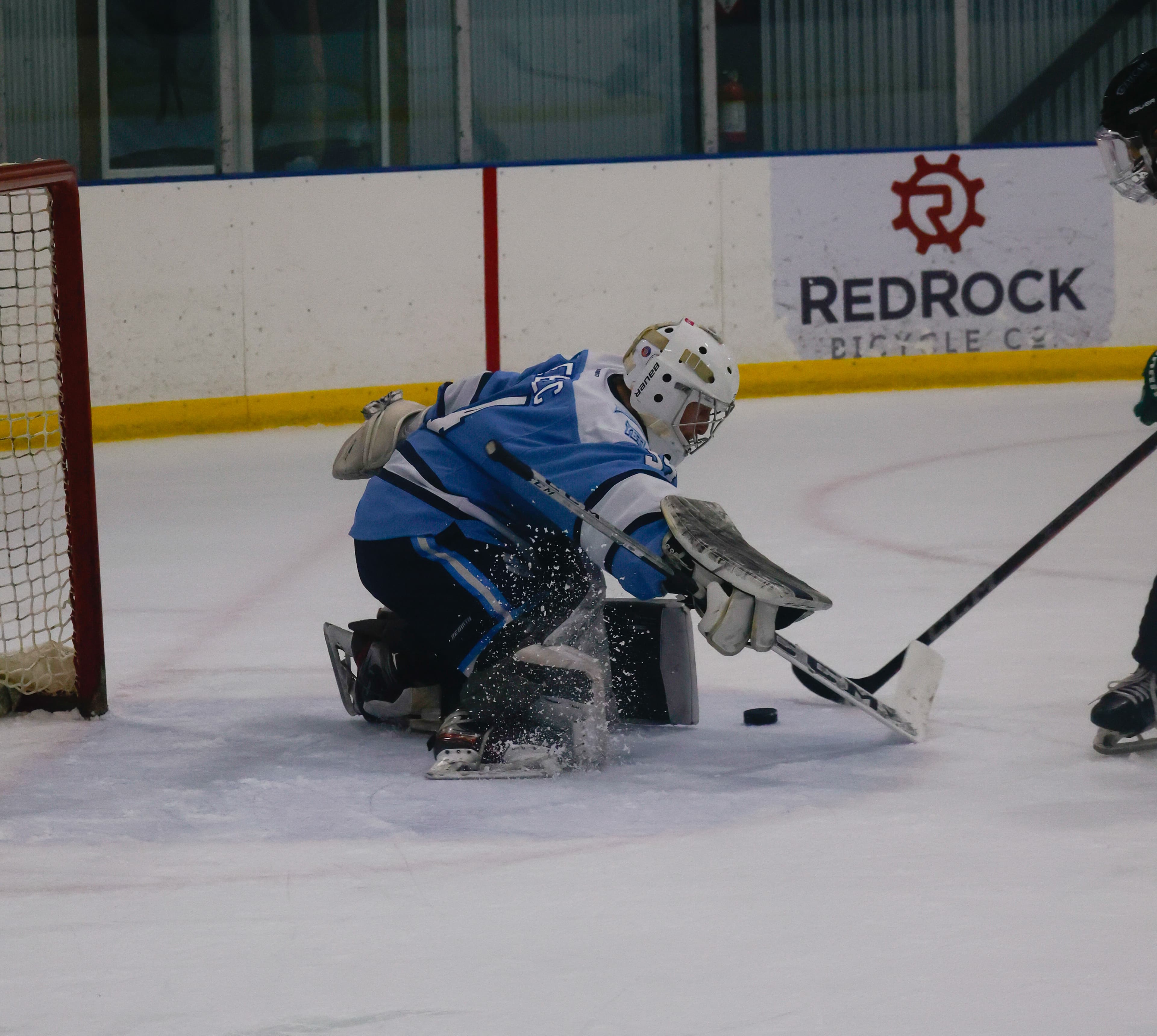 Hockey goalie in blue jersey makes a sliding save with ice spraying near the goal.